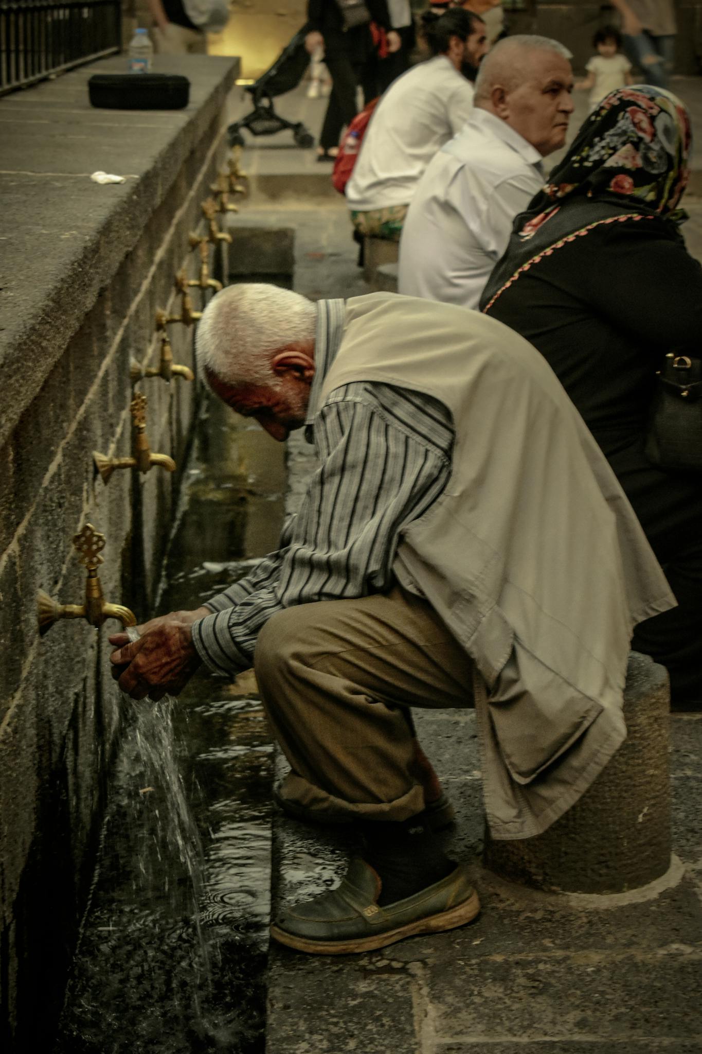 An elderly man washes his hands at a traditional outdoor water fountain.